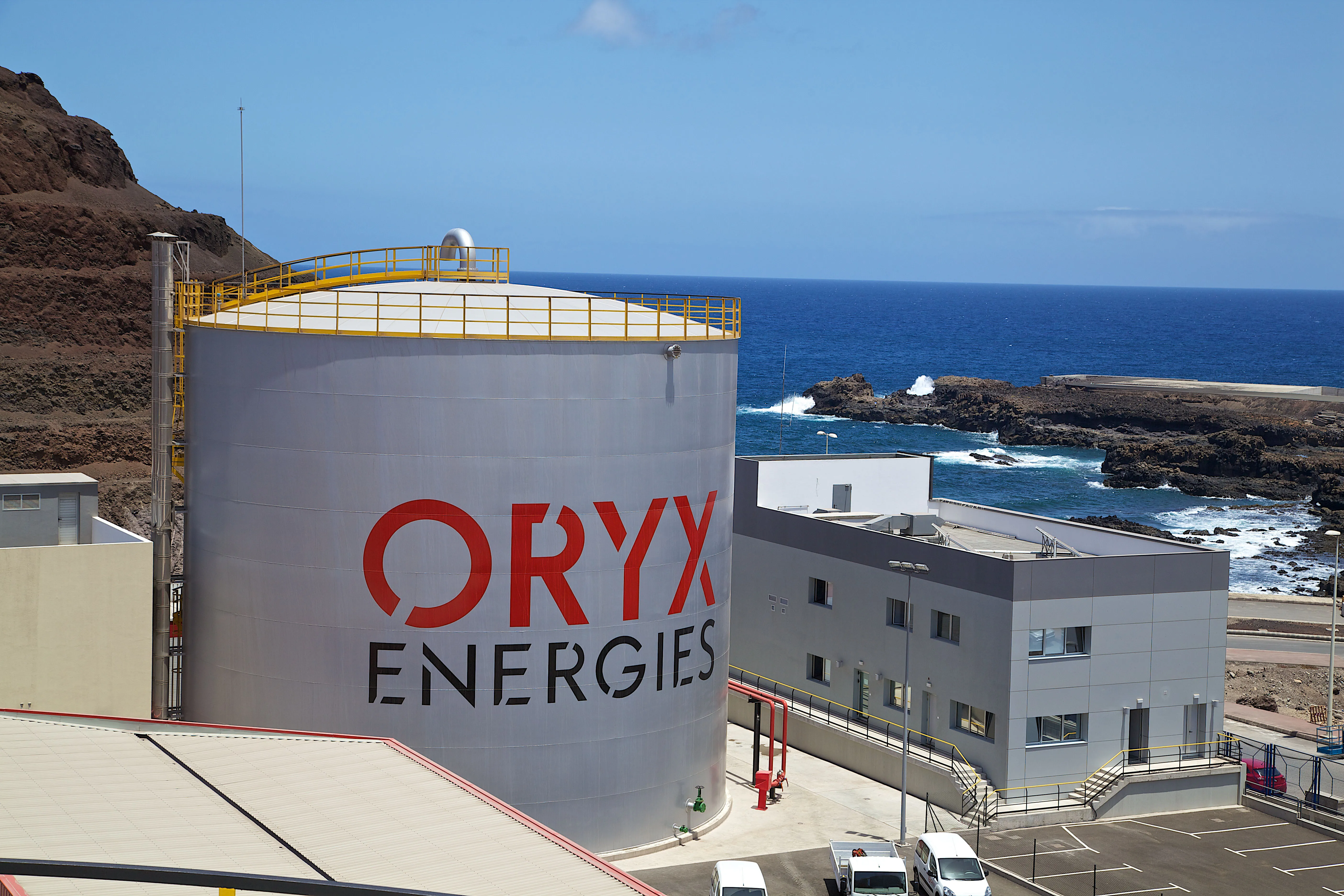 Large ORYX Energies storage tank by the sea, with industrial buildings and rocky coastline.